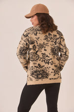Person wearing a beige sweater with black floral pattern and a tan cap on a plain background